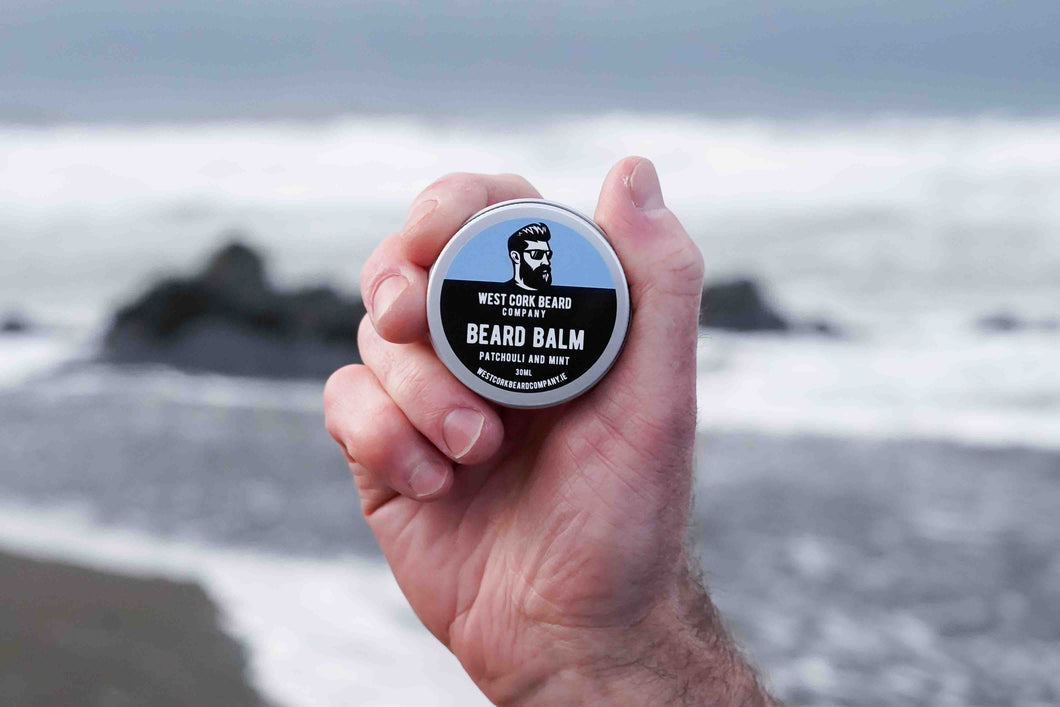 Hand holding a container of West Cork Beard Balm against a beach background