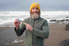 Man on a beach holding a tin of beard balm, pointing towards it