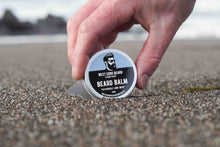 Hand holding a container of West Cork Beard Company beard balm on a sandy surface.