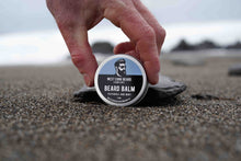 Hand holding a container of West Cork Beard Company beard balm on sand with a blurred background