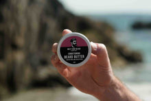 A hand holding a round tin of West Cork Beard Care Conditioning Beard Butter against a blurred outdoor coastal background.