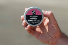 A hand holding a round tin of West Cork Beard Company Conditioning Beard Butter against a blurred beige background.