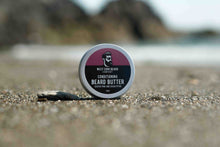 A round tin of West Cork Beard Company Conditioning Beard Butter sits on a sandy beach with a blurred rocky shoreline in the background.