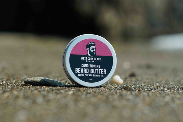 A round tin of West Cork Beard Company Conditioning Beard Butter standing on a sandy surface outdoors, with a pink and black label and a blurred natural background.