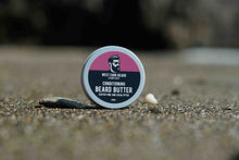 A round tin of West Cork Beard Company Conditioning Beard Butter sits on a sandy surface with a blurred outdoor background.