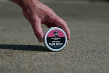 A hand is touching a round tin of West Cork Beard Care Conditioning Beard Butter, which is resting on a textured sandy surface on a beach.