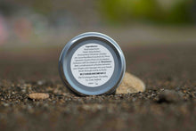 A small round metal tin of West Cork Beard Company Beard butter sits on a textured ground surface with a blurred outdoor background.