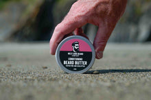 A hand holding a round tin of West Cork Beard Care Conditioning Beard Butter on a textured sandy surface with a blurred outdoor background.
