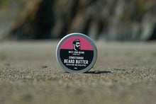A round tin of West Cork Beard Company Conditioning Beard Butter standing upright on a sandy surface with a blurred outdoor background.