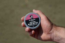 A hand holding a round tin of West Cork Beard Company Beard Butter against a blurred outdoor background.